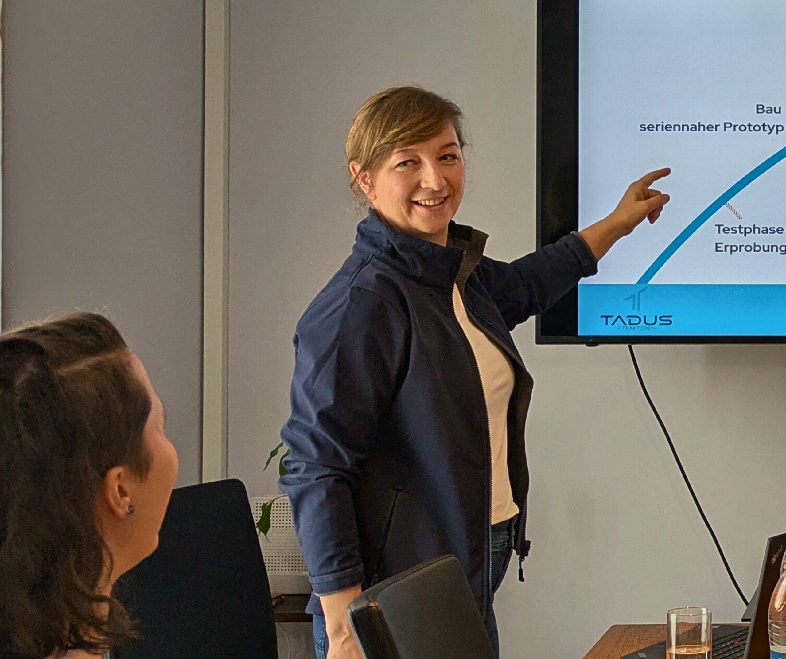 Zu sehen ist die Geschäftsführung von TADUS. Sie zeigt mit der linken Hand auf einen Monitor um etwas zu erklären. Vor ihr sitzt eine weitere Mitarbeiterin des Unternehmens.