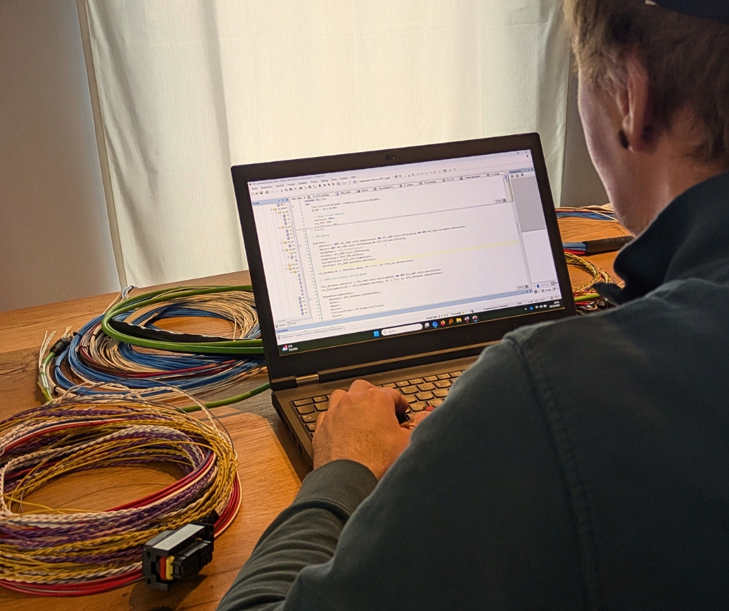 Das Bild zeigt einen Mitarbeiter von TADUS. Er ist Softwareingenieur und programmiert gerade einen Code. Links im Bild sind verschieden Kabelbäume zu sehen.