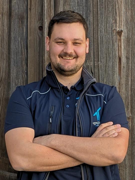 Das Bild zeigt einen Mitarbeiter der TADUS GmbH im Portrait. Der Hintergrund ist eine Holzwand.