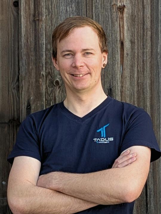Das Bild zeigt einen Mitarbeiter der TADUS GmbH im Portrait. Der Hintergrund ist eine Holzwand.
