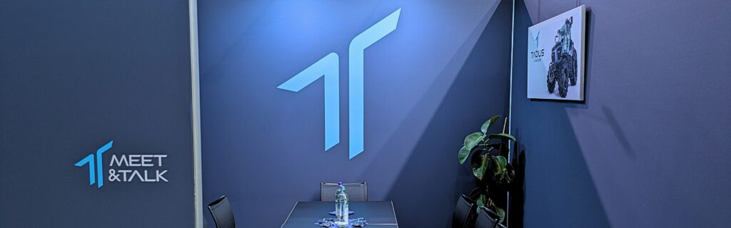 Zu sehen ist ein Ausschnitt des TADUS Messestand der AgriTechnica 2025. Der Raum "Meet&Talk" für Gespräche. Sitzgelegenheiten, ein Tisch, Getränke und Süßigkeiten sowie ein Bild des TADUS T16.20 und einer Zimmerpflanze.
