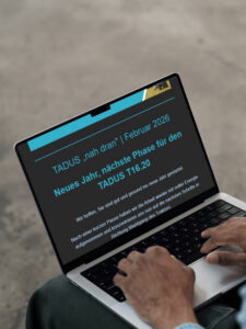 Ein grauer Laptop auf einem Schoss einer Person, die gerade etwas tippt. Der Bildschirm zeigt den Newsletter von TADUS.